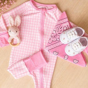 May include: A pink and white gingham baby sleeper with a zipper, a crochet bunny rattle, a pink bandana, and white baby shoes with floral accents. The baby shoes have a size 11 marking.