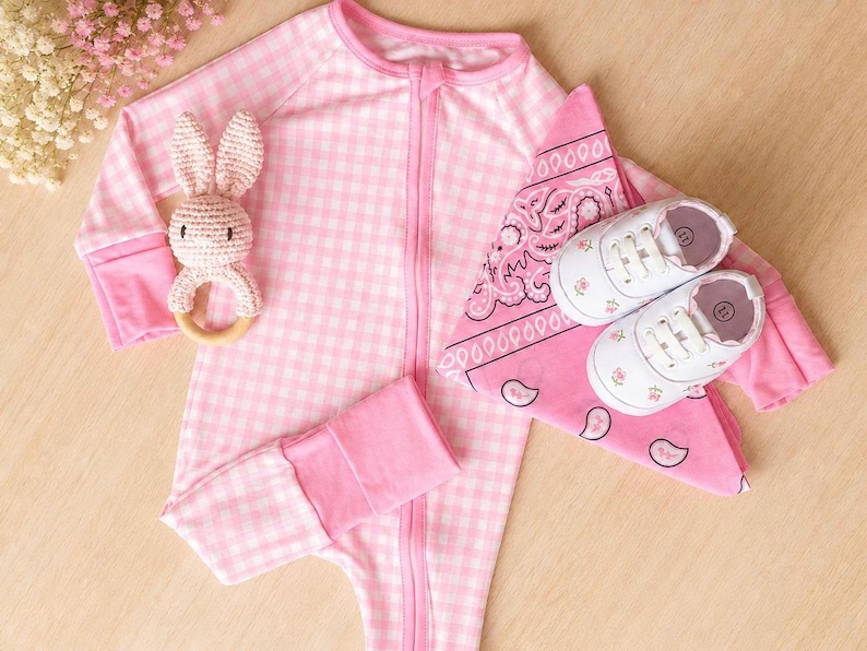 May include: A pink and white gingham baby sleeper with a zipper, a crochet bunny rattle, a pink bandana, and white baby shoes with floral accents. The baby shoes have a size 11 marking.