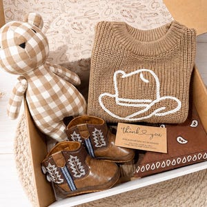 Op de afbeelding: Een geschenkdoos met een beige en wit geblokte teddybeer, een beige gebreide trui met een witte omtrek van een cowboyhoed, bruine kunstleren cowboylaarzen met witte stiksels en een bruine bandana met witte patronen en een bedankkaartje.