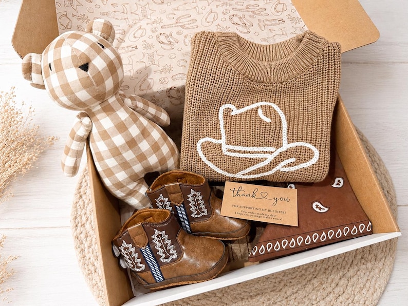 Op de afbeelding: Een geschenkdoos met een beige en wit geblokte teddybeer, een beige gebreide trui met een witte omtrek van een cowboyhoed, bruine kunstleren cowboylaarzen met witte stiksels en een bruine bandana met witte patronen en een bedankkaartje.