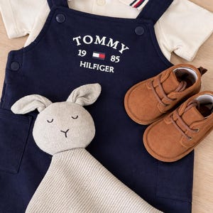 A navy blue baby outfit set with a white collared shirt, navy blue overalls with "TOMMY HILFIGER" text, brown suede baby shoes, and a grey bunny comforter. The set is displayed on a light wood surface.