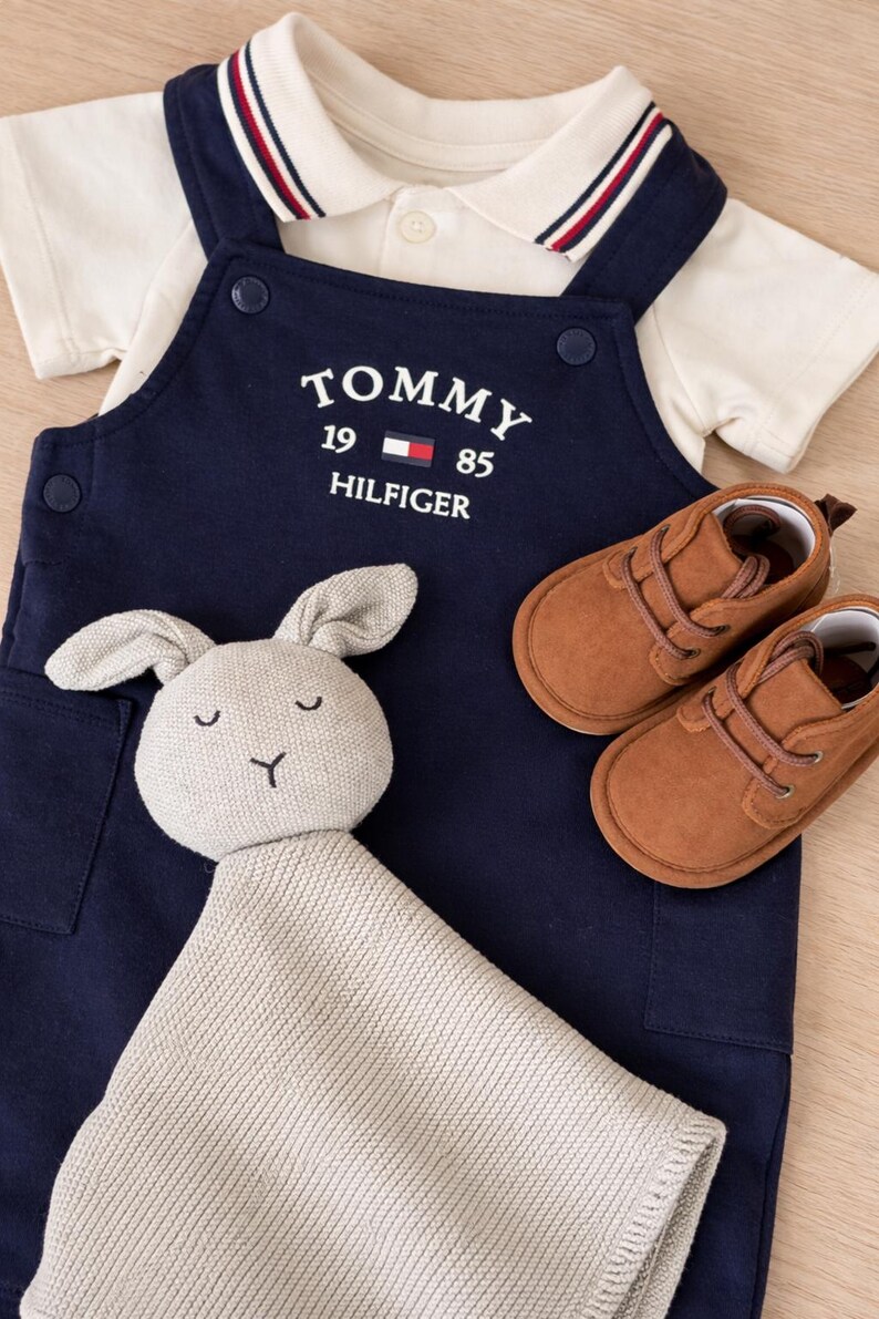 A navy blue baby outfit set with a white collared shirt, navy blue overalls with "TOMMY HILFIGER" text, brown suede baby shoes, and a grey bunny comforter. The set is displayed on a light wood surface.
