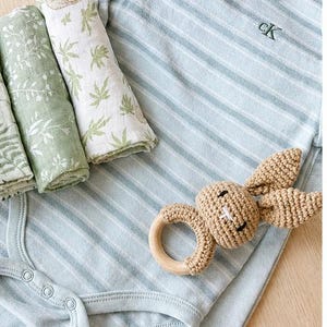May include: A baby clothing set featuring a light blue striped onesie with the embroidered initials "cK", and a crochet bunny rattle with a wooden ring. Rolled muslin cloths with green leaf patterns are also included.