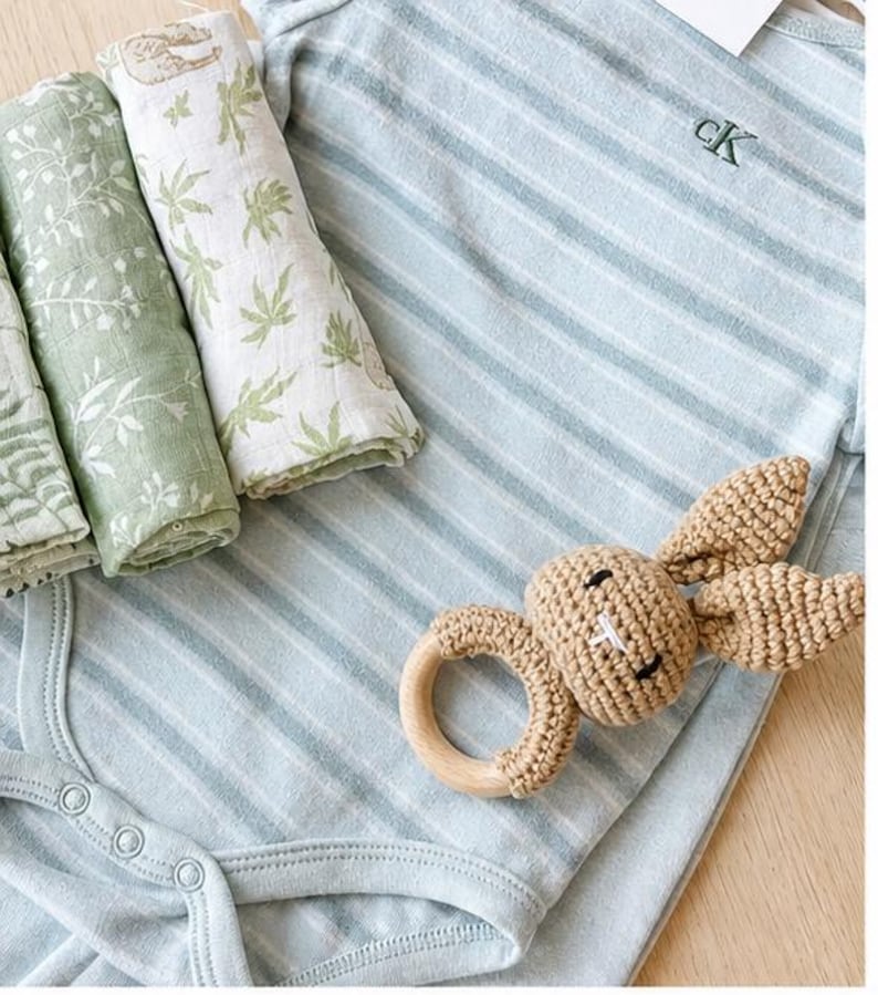May include: A baby clothing set featuring a light blue striped onesie with the embroidered initials "cK", and a crochet bunny rattle with a wooden ring. Rolled muslin cloths with green leaf patterns are also included.