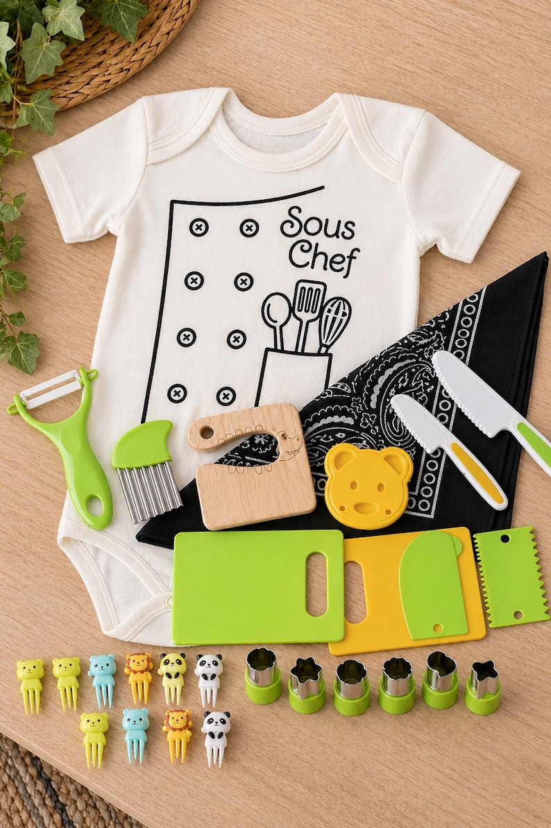 Puede incluir: Un body blanco para beb&eacute; con un gr&aacute;fico de "Sous Chef", junto con una variedad de utensilios de cocina, incluyendo un pelador, cuchillos, tablas de cortar y pinchos para alimentos. El conjunto incluye cortadores de galletas con forma de oso y una bandana negra.