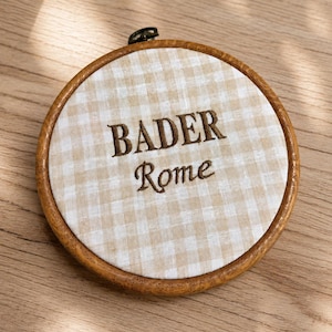 May include: A round embroidery hoop with a light brown wooden frame. The fabric inside has a beige and white checkered pattern. The words "BADER" and "Rome" are embroidered in brown thread.