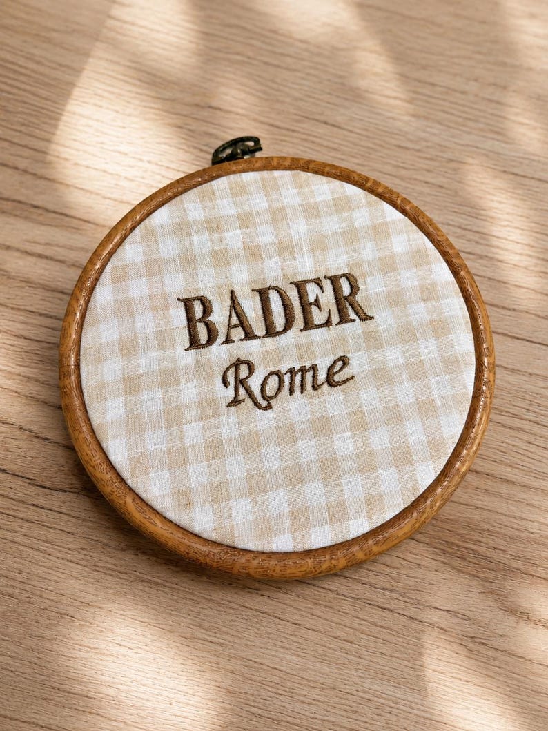 May include: A round embroidery hoop with a light brown wooden frame. The fabric inside has a beige and white checkered pattern. The words "BADER" and "Rome" are embroidered in brown thread.