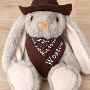 Op de afbeelding: Een pluche konijnknuffel met zachte lichtbruine vacht, voorzien van een donkerbruine vilten cowboyhoed en een donkerbruine bandana met een wit paisley-patroon en de naam Weston in witte schrijfletters op de voorzijde geborduurd.