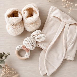 May include: A set of baby items, including cream-colored plush booties, a crocheted bunny rattle with a wooden ring, and a matching bunny ear bonnet. The items are arranged on a light wooden surface, creating a soft and neutral aesthetic.