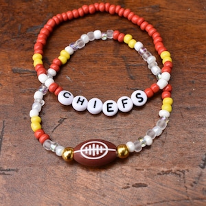 May include: Two beaded bracelets featuring red, yellow, and clear beads. One bracelet displays white letter beads spelling CHIEFS. The other bracelet includes a brown football-shaped bead with white laces, flanked by two metallic gold beads.