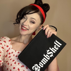 Puede incluir: Clutch negra con la palabra "Bombshell" en letras góticas blancas. El clutch tiene un asa negra. La persona de la imagen lleva una diadema roja y una blusa blanca con diseños de fresas rojas.