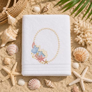 May include: A white rectangular bath towel featuring an embroidered oval frame made of pearls, accented at the bottom with a cluster of blue seashells, a pink starfish, and a small gold shell. The towel has three horizontal textured bands.