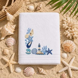 Könnte beinhalten: Ein weißes, rechteckiges Badetuch mit einer gestickten Küstenlandschaft in der unteren Ecke. Die Stickerei zeigt einen Leuchtturm, blaue Seesterne, verschiedene Muscheln und Korallenzweige in Blau-, Beige- und Weißtönen.