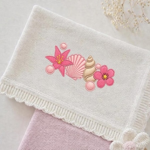 May include: A white knitted baby blanket with a scalloped edge and embroidered seashell and flower design in shades of pink and tan. The blanket is folded over a pink blanket.