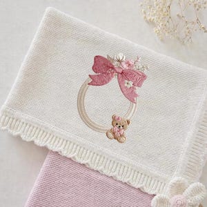 May include: A white knitted baby blanket with a pink embroidered bow and teddy bear design. The blanket has a scalloped edge and a pink blanket is visible underneath.
