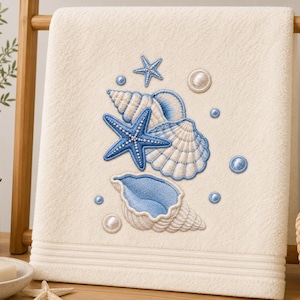 Könnte beinhalten: Ein cremefarbenes Handtuch mit einer Stickerei aus blauen und weißen Muscheln, Seesternen und kleinen kreisförmigen Akzenten. Die Stickerei ist mittig auf dem Handtuch platziert, das eine strukturierte Stoffoberfläche und horizontale Linien aufweist.