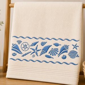 May include: A cream-colored bath towel featuring a horizontal band of embroidered blue seashells, starfish, and sand dollars. The design is framed by two wavy blue lines above and below the marine motifs, with subtle horizontal ribbed texture.