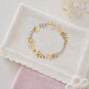 May include: A white knitted baby blanket featuring an embroidered wreath of seashells, starfish, and foliage in gold and grey. The blanket has a scalloped edge and covers a pink blanket.