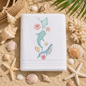 Puede incluir: Una toalla rectangular blanca con un diseño bordado de una ballena azul claro, un tiburón ballena verde azulado, una raya verde claro, una estrella de mar rosa y dos flores de hibisco rosas, con pequeñas conchas marinas dispersas.