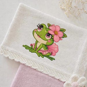 May include: White knitted baby blanket featuring a cartoon frog wearing a pink tutu and a flower crown. The frog is green with pink accents. The blanket has a scalloped edge and is folded over a pink blanket.