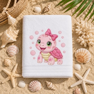 Puede incluir: Una toalla rectangular blanca con un gráfico bordado de una tortuga de dibujos animados rosa con un lazo rosa en la cabeza, rodeada de pequeñas burbujas rosas. La tortuga tiene grandes ojos verdes y un vientre amarillo, centrada en la tela.