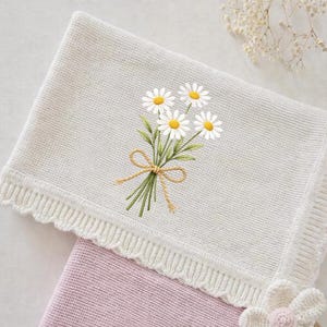 May include: A white knitted blanket with a daisy bouquet embroidery. The bouquet features white daisies with yellow centers and green leaves, tied with a brown twine bow. The blanket has a scalloped edge and is partially covering a pink blanket.