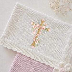 Może przedstawiać: Kremowy dzianinowy kocyk dla dziecka ze złotym krzyżem wyszywanym różowymi kwiatami i różową kokardą. Kocyk ma falisty brzeg i częściowo zakrywa różowy kocyk.