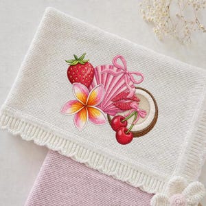 May include: White knitted blanket with a decorative embroidered design featuring a strawberry, plumeria flower, pink seashell, lips, cherries, and a coconut. The blanket has a scalloped edge.