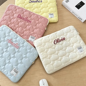 May include: Four quilted laptop sleeves in pastel colors: yellow, pink, light blue, and cream. Each sleeve features a cloud pattern and a name embroidered in a contrasting color. The names are "Sauber", "Deariloon", "Amelia", and "Olivia".