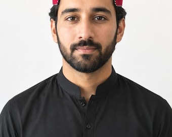 May include: A red, embroidered cap with a white and black trim, worn by a person wearing a black button-down shirt. The cap is a traditional style, likely for cultural or religious purposes.