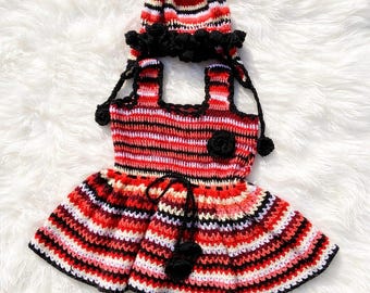 May include: A handmade crochet baby dress and matching hat set. The dress features a red, black, and white striped pattern with a black flower accent. The hat has a similar striped design and black pom-pom trim. The set is designed for a baby.