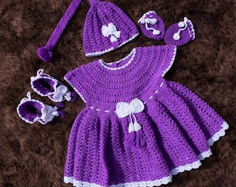 May include: A handmade purple crochet baby dress set with a matching hat, booties, and mittens. The dress features a white bow and tassel detail. The set is trimmed with white crochet edging and is displayed on a brown surface.