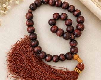 May include: A string of dark brown, spherical wooden prayer beads arranged in a circular pattern. The beads feature a visible wood grain texture and are connected by a cord that terminates in a large, dense, rust-colored thread tassel.