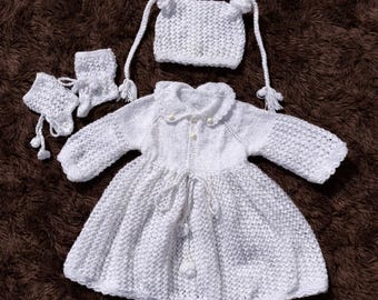 May include: A white knitted baby dress set, including a dress, hat, mittens, and booties. The dress features a Peter Pan collar and button closure. The hat has ear-like details and ties. The set is displayed on a brown surface.