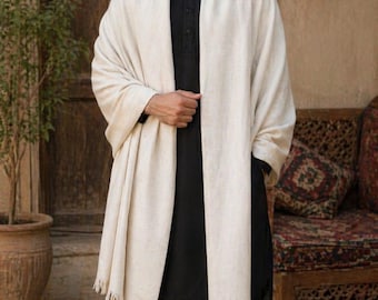 May include: A person wearing a black outfit with a cream-colored shawl. The shawl has a fringed edge. The person is standing in front of a wooden door and a decorative bench with a patterned cushion.
