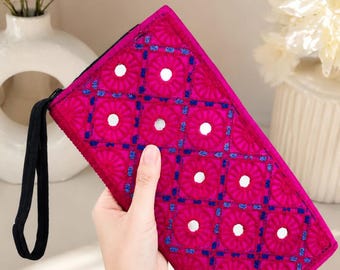 May include: A rectangular fuchsia clutch bag featuring a grid pattern of embroidered floral motifs in dark blue thread, accented with small circular mirrors in the center of each square. The clutch includes a black wrist strap and a zipper closure.