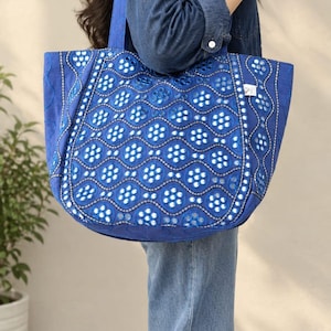 Peut inclure: Un grand sac fourre-tout bleu orné d'un motif géométrique répétitif composé de découpes circulaires et d'accents réfléchissants argentés. Le sac possède deux anses assorties et une base large et structurée avec des broderies détaillées.