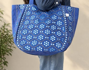 May include: A large blue tote bag featuring a repeating geometric pattern of circular cutouts and silver-toned reflective accents. The bag has two matching blue shoulder straps and a structured, wide base, showcasing intricate embroidery details.