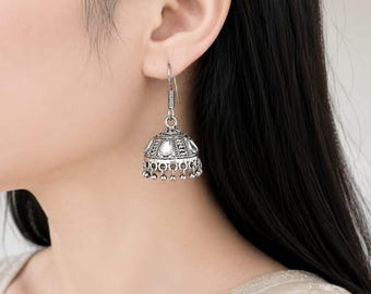 May include: A silver-toned jhumka earring featuring an oxidized metal finish, intricate embossed patterns, and small white teardrop-shaped inlays. The bell-shaped design is accented with a row of tiny dangling silver beads along the bottom edge.