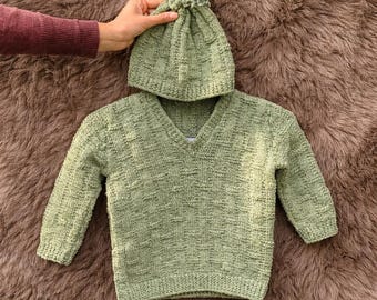 May include: A light green knitted baby sweater and matching beanie with a pom-pom. The sweater has a V-neck and long sleeves. The set is made of a textured knit fabric.