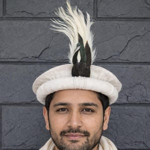 Puede incluir: Un sombrero pakol de lana color crema con un borde enrollado y un adorno de plumas. La pluma es principalmente blanca con puntas de color marrón oscuro y verde iridiscente, colocada en posición vertical en el centro frontal de la copa.