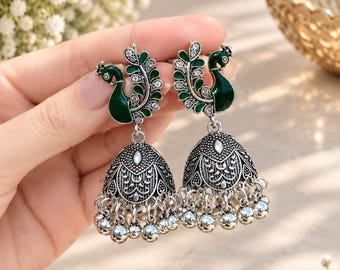 May include: A pair of silver-toned jhumka earrings featuring a dark green peacock motif at the top. The peacock design is accented with small clear stones. The bell-shaped base has intricate embossed patterns and a row of small silver-toned beads.