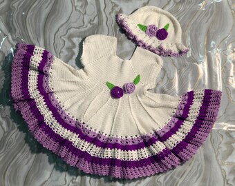 May include: A handmade crochet baby dress and matching hat set. The dress is white with purple and white striped trim and purple flower accents. The hat is white with a purple flower and green leaf accents. The set is designed for a baby.