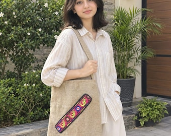 May include: A beige textured tote bag featuring a single diagonal strap across the front. The strap is decorated with a vibrant, multi-colored embroidered pattern in shades of pink, yellow, blue, and black, set against a neutral fabric base.