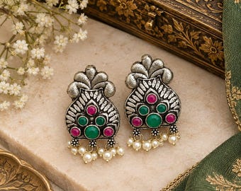 May include: A pair of oxidized silver-tone stud earrings featuring a fan-shaped top, central green and pink cabochon stones, and small dangling white faux pearls. The metal has a textured, darkened finish with intricate engraved detailing.