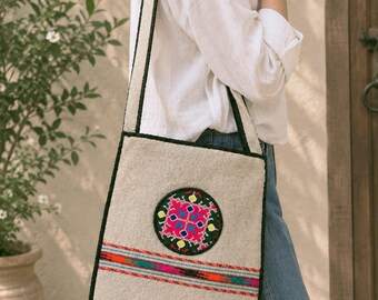 May include: A beige rectangular shoulder bag with black trim, featuring a circular patch with colorful pink, green, and blue embroidery. A horizontal band of multi-colored woven patterns sits below the patch, and the bottom edge has fringe.