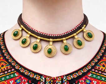 May include: A gold-toned necklace with seven circular pendants, each featuring a green stone. The necklace has a brown cord with red and white accents. The person is wearing a colorful embroidered top.