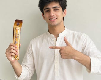 May include: A long, cylindrical food item packaged in a clear plastic sleeve with a brown and gold label. The packaging features decorative patterns and text in a non-Latin script. The item is light tan in color with a textured, granular surface.