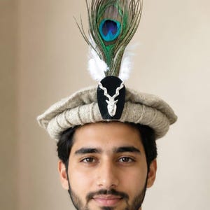 Può includere: Un cappello pakol in lana beige con una toppa centrale nera decorata con un emblema in metallo argentato a forma di testa di cervo. Il cappello è ornato da una piuma di pavone e due piccole piume bianche rivolte verso l'alto.