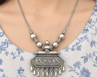May include: A silver-toned metal necklace featuring a large, rectangular pendant with intricate floral engravings and a row of small, dangling teardrop-shaped charms. The chain is composed of smooth, round beads and a sturdy, polished metal snake chain.